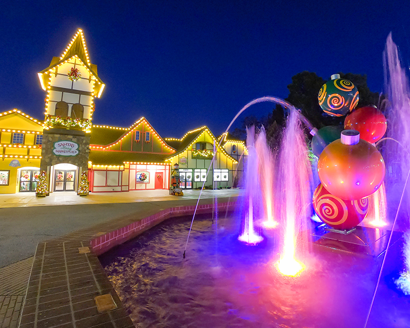 The outside of Santa's Merry Marketplace at night lit with lights along the top of the building. A Christmas ornament fountain is in the foreground and lit by colorful lights in the water.